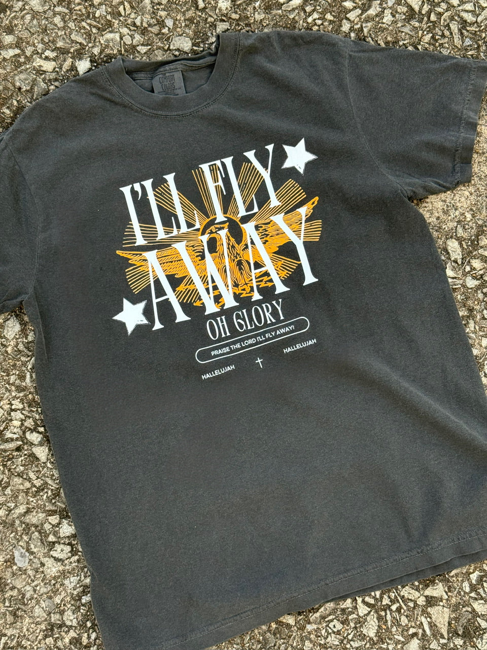Front of black/dark grey short sleeve t-shirt with the text "I'll fly away, Oh Glory." printed in white in front of a graphic of a gold vintage eagle with sun rays and two white stars. The shirt is laying on the ground.