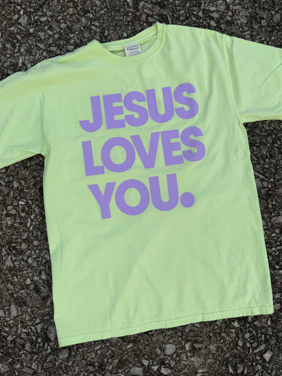 Front of neon yellow short sleeve t-shirt with "Jesus loves you" printed in bright lilac purple. The shirt is laying on the ground.