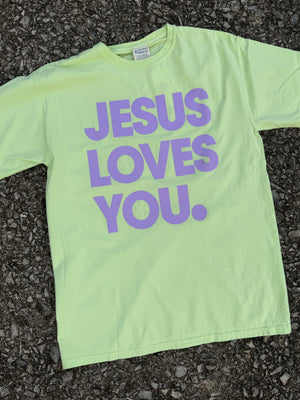 Front of neon yellow short sleeve t-shirt with "Jesus loves you" printed in bright lilac purple. The shirt is laying on the ground.
