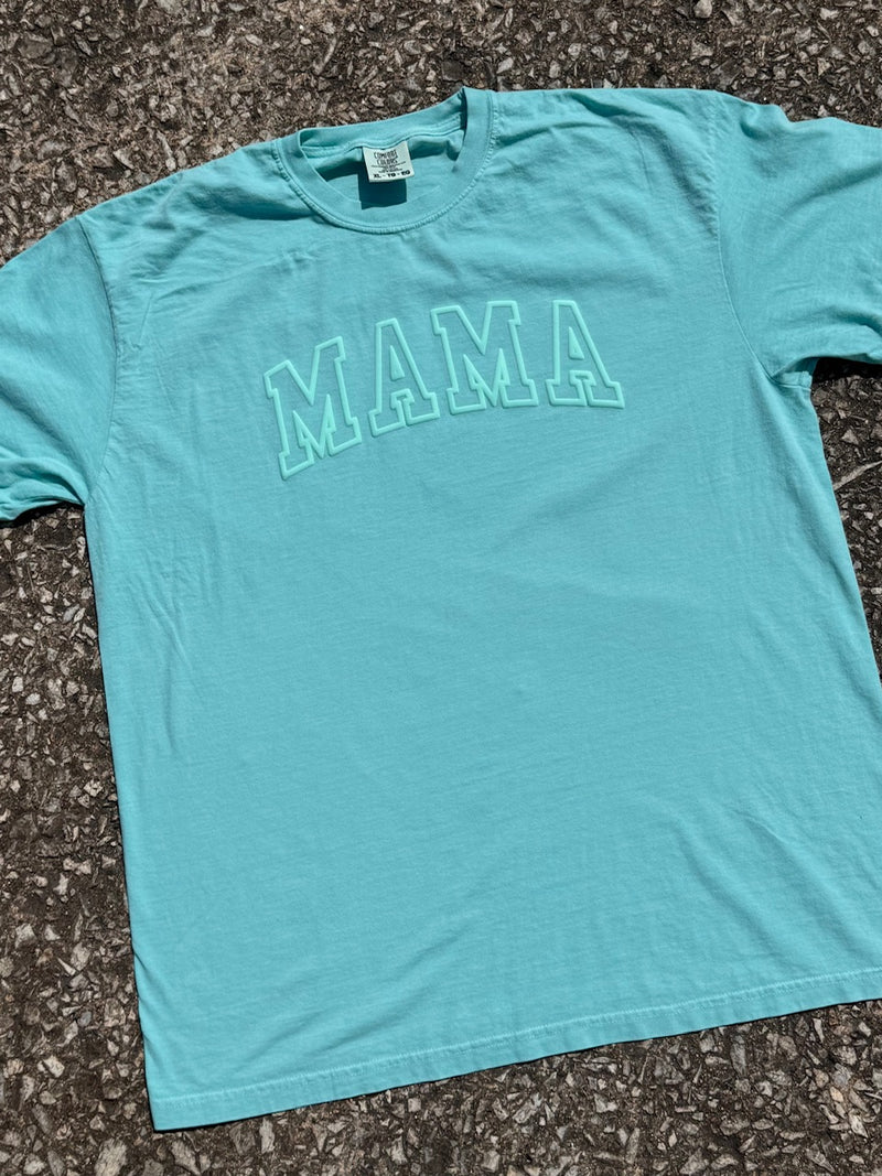 Front of Turquoise short sleeve t-shirt with 'MAMA' text printed in a light turquoise in puffy ink. The shirt is laying on the floor.
