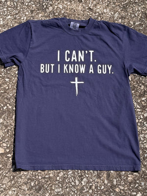Front of navy blue short sleeve T-shirt with "I can't. but I know a guy." printed in cream with a small simple cross graphic below the text. The shirt is laying on the floor.