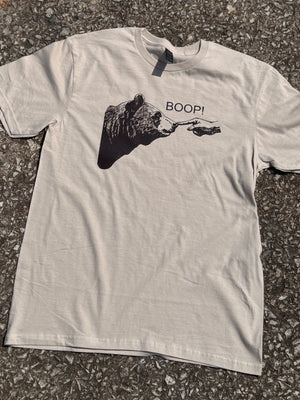 Front of Heather Sand short sleeve T-shirt with a graphic of a bear with a hand poking the bear's nose with the text "Boop!" printed in brown. The shirt is laying on the floor.