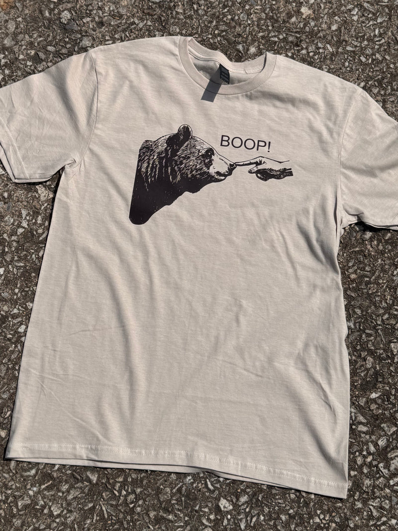 Front of Heather Sand short sleeve T-shirt with a graphic of a bear with a hand poking the bear's nose with the text "Boop!" printed in brown. The shirt is laying on the floor.