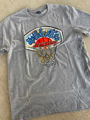 A heather gray short-sleeve t-shirt laid flat on a wood floor, featuring a large graphic print of a basketball hoop, an orange basketball, and the word 'Wildcats' in blue script.