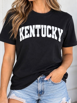 A woman wearing a black short-sleeve t-shirt with 'KENTUCKY' printed in large white varsity-style letters across the chest, paired with light-wash distressed denim shorts.