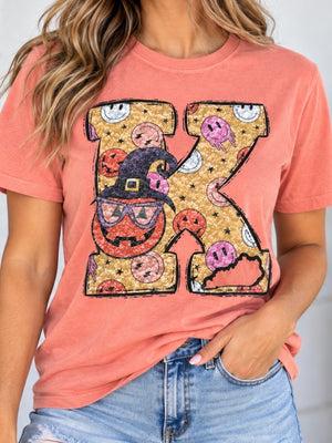 A woman wearing a coral pink t-shirt with a large glittery letter 'K' graphic featuring Halloween-themed illustrations like a witch hat, jack-o'-lantern, and ghosts, paired with blue jeans.