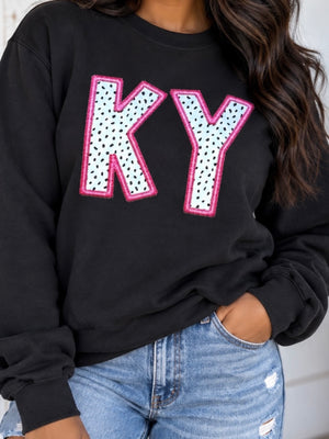 A person wearing a black crewneck sweatshirt with large decorative letters 'KY' outlined in pink and filled with a white polka-dot pattern, paired with light blue jeans.
