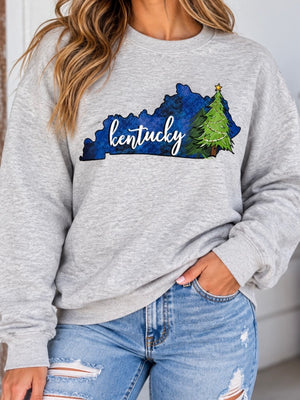 A woman wearing a heather gray crewneck sweatshirt with a graphic of the state outline of Kentucky in blue watercolor, decorated with a green Christmas tree and the word 'kentucky' in white script. She is wearing light-wash distressed blue jeans.