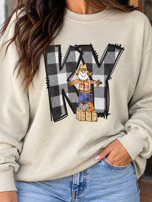A woman wearing a beige crewneck sweatshirt with a graphic print of the letters 'KY' in a black and white checkered pattern, featuring a cartoon scarecrow standing in front of it, paired with blue jeans.
