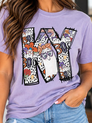 A woman wearing a lavender purple graphic t-shirt with a large 'KY' design featuring a ghost wearing purple glasses and other whimsical halloween doodles, paired with blue jeans.