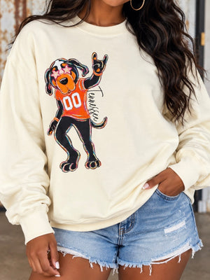 A woman wearing a cream-colored crewneck sweatshirt with a cartoon graphic of a dog dressed in an orange football jersey, paired with distressed denim shorts.