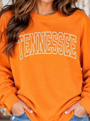 A woman wearing an orange crewneck sweatshirt with 'TENNESSEE' printed in white arched block letters across the chest, paired with blue jeans.