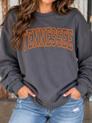 A woman wearing a dark charcoal gray sweatshirt with 'TENNESSEE' printed across the chest in orange varsity-style block letters, paired with light blue jeans.
