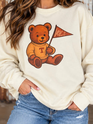 A woman wearing a cream-colored crewneck sweatshirt with a graphic of a brown teddy bear holding a flag that says 'VOL'S' and wearing a sweater with a 'T' on the chest. She is also wearing blue jeans.