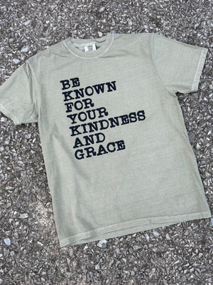 Front of greenish-gray short sleeve crew neck t-shirt with the text "Be known for your kindness and grace" in a typewriter font printed in black. The shirt is laying flat on the floor.