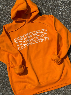 Front of bright Orange long sleeve hoodie with 'TENNESSEE' printed on it in white, laid flat on a textured surface.