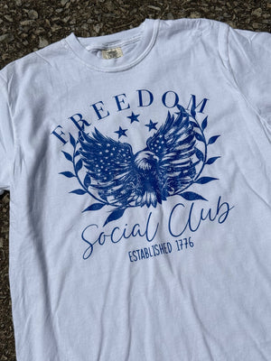 Front of light blue short sleeve t-shirt with a graphic of a bald eagle with three stars above it and the text "Freedom" above the eagle and the text "Social club, Established 1776" below the eagle printed in dark blue. The shirt is laying flat on the floor.