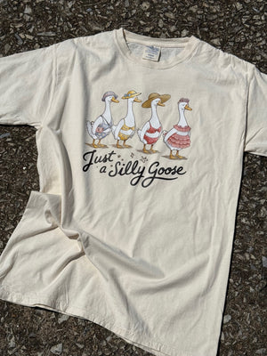Front of Beige short sleeve t-shirt with cartoon geese wearing swimsuits and text 'Just a Silly Goose'  printed on the shirt in black. The shirt is laying flat on the floor.