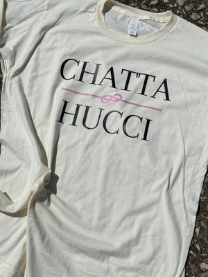 Front of cream colored t-shirt with the text "Chatta Hucci" printed in black with a pink sailors rope in the middle printed in pink. The shirt is laying flat on the floor.