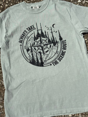 Front of light green short sleeve t-shirt with graphic of a compass with pine trees and mountains inside it and the text "Always Take the scenic route." around the outside of the compass printed in black. The shirt is laying flat on the floor.
