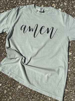 Front of light sage green short sleeve t-shirt with the text "amen" in a cursive font printed in black across the chest. The shirt is laying flat on the floor.