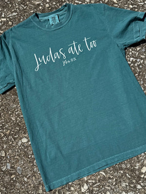 Front of teal short sleeve t-shirt with the text "Judas ate too John 13:26" in a cursive font printed in white across the chest. The shirt is laying flat on the floor.