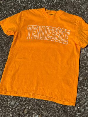 Front of bright orange short sleeve t-shirt with the outline of the text "Tennessee" printed in white across the chest in white. The shirt is laying flat on the floor.