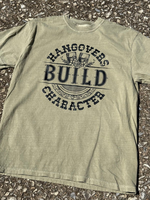 Front of grungy green short sleeve t-shirt with the text "Hangovers build character" in a western font with a graphic of two whiskey glasses with flowers around them printed in black. The shirt is laying flat on the floor.