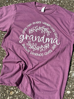 Front of heather plum purple short sleeve t-shirt with the text "grandma" then two outlines of vines forming a small circle around the text "grandma" then the text "Keeps faith, Shares wisdom, lives love, shows patience, gives joy." forming another circle around that printed in white. The shirt is laying flat on the floor.