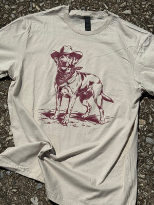 Front of Beige short sleeve t-shirt with a graphic of a labrador retriever dog wearing a cowboy hat and bandana  printed in dark red. The shirt is laying flat on the floor.