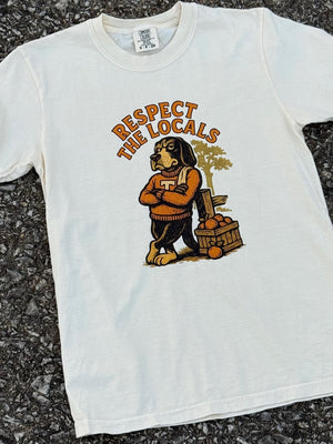 Front of white short sleeve T-shirt with a graphic of a hound dog wearing an orange sweater leaning against a fence post and a crate of oranges with the text "respect the locals" above printed in orange. The shirt is laying on the floor.