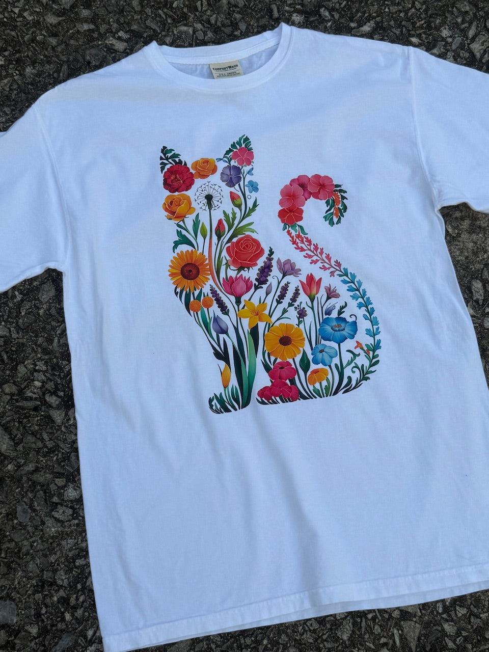 Front of white short sleeve T-shirt with graphic of an outline of a cat filled in with colorful flowers." The shirt is laying on the floor.