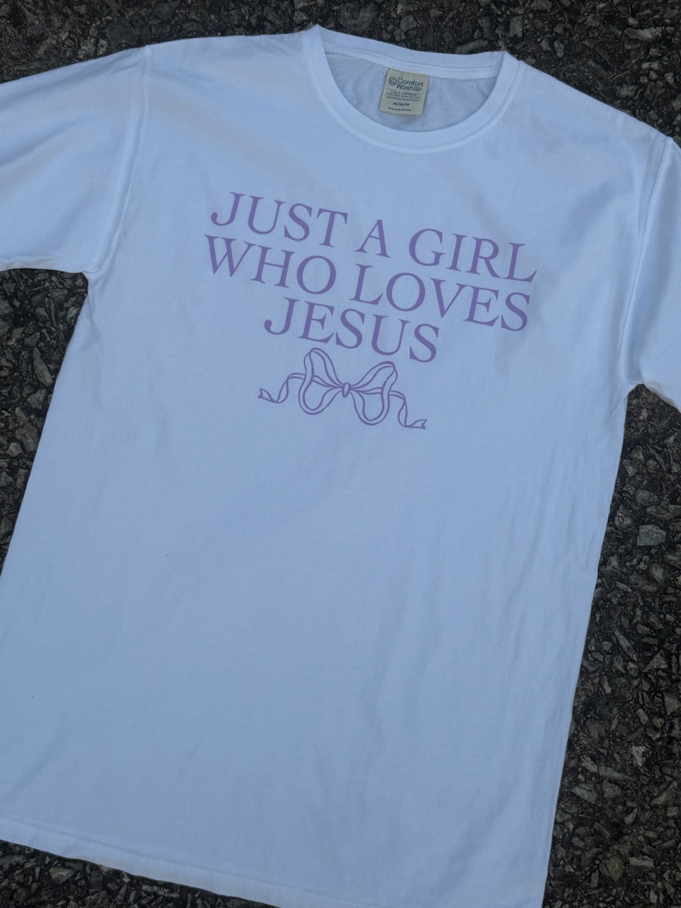 Front of a short sleeve white T-shirt with the text "Just a girl who loves jesus" and a bow outline graphic is underneath it printed in purple. The shirt is laying on the floor.