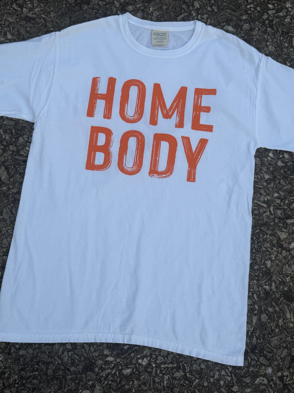 Front of white short sleeve T-Shirt with the text "HOME BODY" printed in orange. The shirt is laying on the floor.