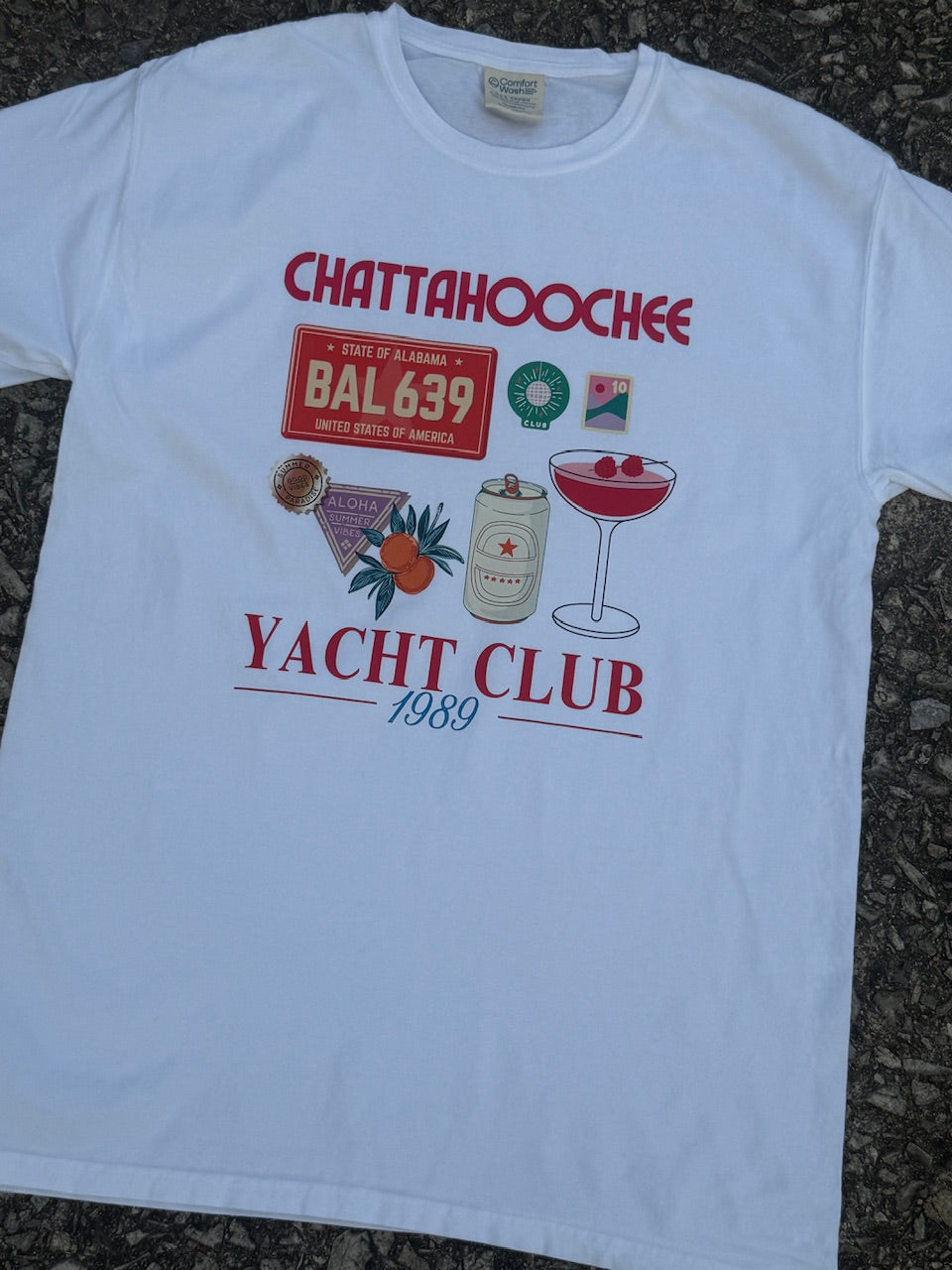 Front of white short sleeve T-shirt with the text "Chattahoochee Yacht Club" printed in red, the text "1989" printed in blue, with the graphics of an Alabama license plate, cocktail, soda can, disco ball, post stamp, oranges, and a purple sign that says "Aloha summer vibes". The shirt is laying on the ground.
