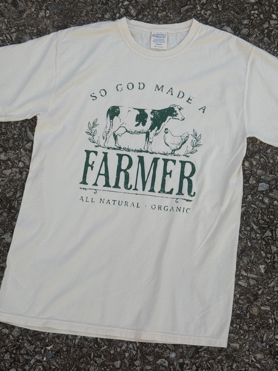 Front of Cream short sleeve T-shirt with the text "So God made a FARMER, all natural, organic." with a dairy cow and a chicken graphic printed in dark green. The shirt is laying on the floor.