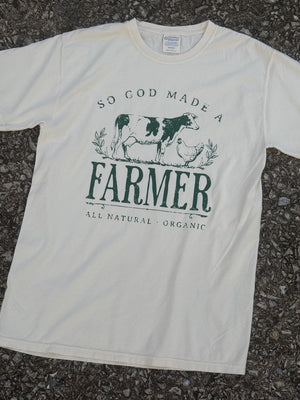 Front of Cream short sleeve T-shirt with the text "So God made a FARMER, all natural, organic." with a dairy cow and a chicken graphic printed in dark green. The shirt is laying on the floor.