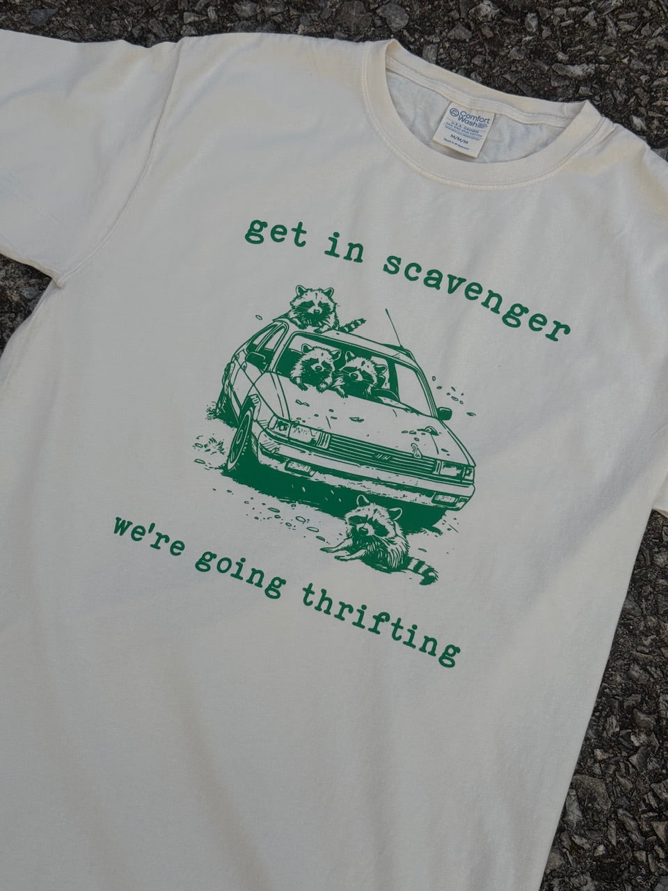 Front of short sleeve cream T-shirt with a graphic of raccoons in a car with the text "get in scavenger" above and "we're going thrifting" below printed in green. The shirt is laying on the floor.