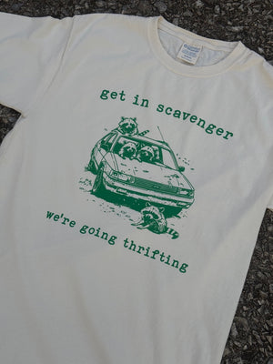 Front of short sleeve cream T-shirt with a graphic of raccoons in a car with the text "get in scavenger" above and "we're going thrifting" below printed in green. The shirt is laying on the floor.