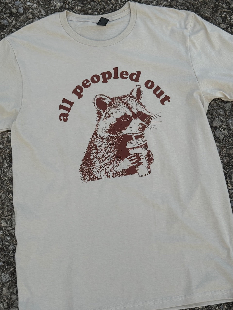 Front of short sleeve heather sand t-shirt with the text "all peopled out" with a graphic of a raccoon drinking out of a coffee cup printed in brown. The shirt is laying on the floor.