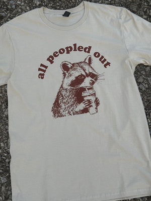 Front of short sleeve heather sand t-shirt with the text "all peopled out" with a graphic of a raccoon drinking out of a coffee cup printed in brown. The shirt is laying on the floor.