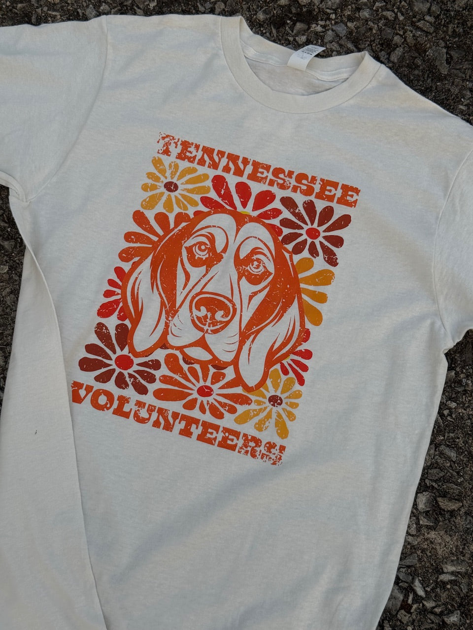 Front of short sleeve heather off white t-shirt with an orange outline of a hound dog's face with vintage flower designs that are orange, yellow, and brown with orange text "Tennessee" above and "Volunteers!" below printed on the shirt. The shirt is laying on the floor.