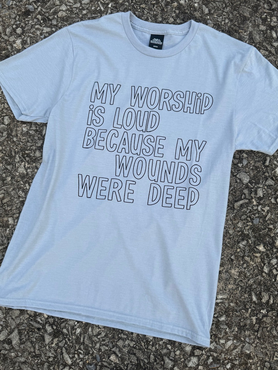 Front of short sleeve heather white t-shirt with the text "My worship is loud because my wounds were deep" printed in black in an outline font. The shirt is laying on the floor.