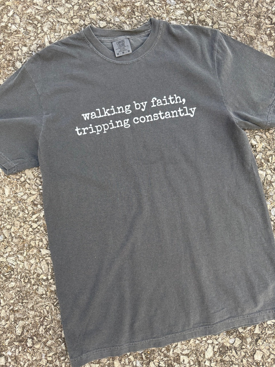Front of short sleeve Gray t-shirt with white text 'walking by faith, tripping constantly' printed on it. The shirt is laying on the floor.