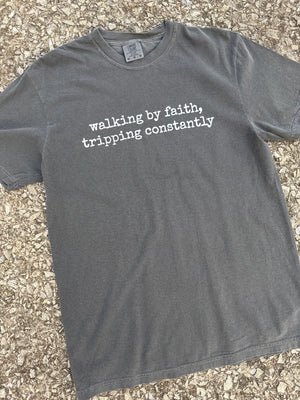 Front of short sleeve Gray t-shirt with white text 'walking by faith, tripping constantly' printed on it. The shirt is laying on the floor.