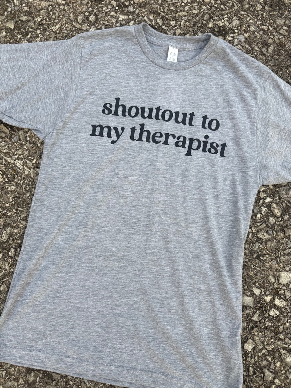 Front of short sleeve heather gray T-shirt with the text "shoutout to my therapist" printed in black. The shirt is laying on the floor.