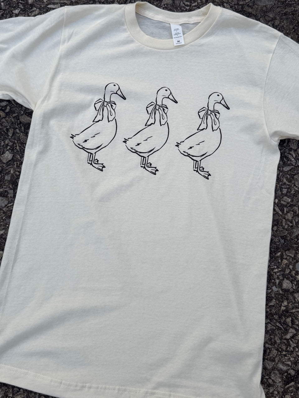 Front of short sleeve white t-shirt with a graphic of the outline of three geese with bows around their necks standing in a row printed in black. The shirt is laying on the floor.