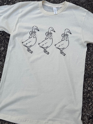 Front of short sleeve white t-shirt with a graphic of the outline of three geese with bows around their necks standing in a row printed in black. The shirt is laying on the floor.