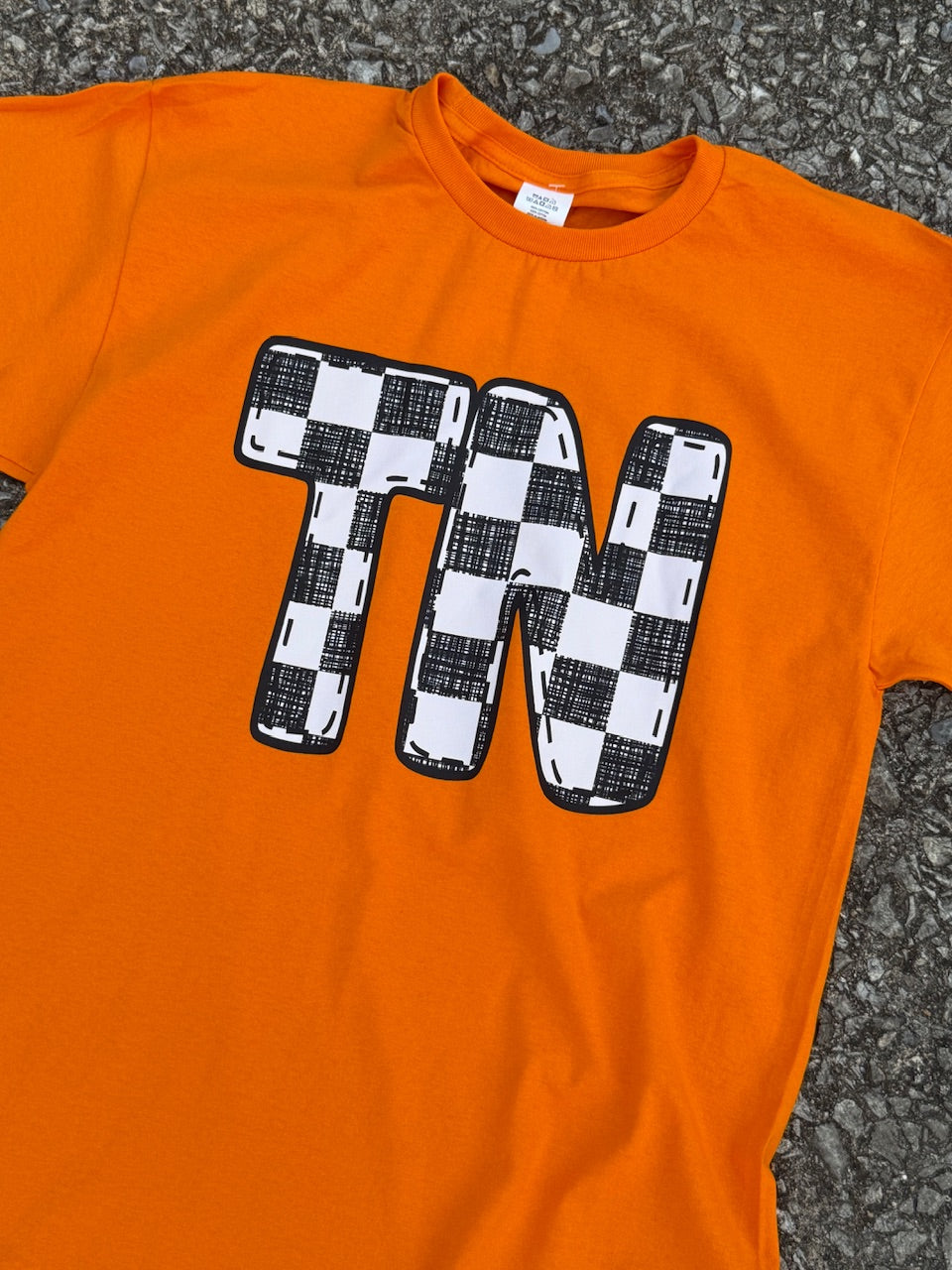 Front of short sleeve bright orange shirt with a large outline of the letters TN with black and white checkerboard filling it in, printed on the shirt. The shirt is laying on the floor.