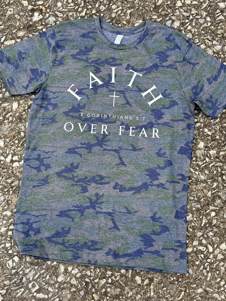 Green Camouflage t-shirt with the text 'Faith Over Fear' and a small cross graphic  is printed on the shirt in white. It is laying on a textured surface.